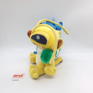 Puede incluir: Un peluche de una criatura amarilla con antenas largas, un parche ocular azul oscuro y una mancha verde en el costado, colocado frente a un peluche azul. Una peque&ntilde;a etiqueta cuadrada blanca con el texto rojo KPTD y A0608 es visible.