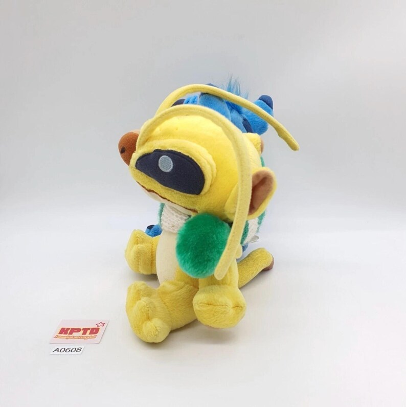 Puede incluir: Un peluche de una criatura amarilla con antenas largas, un parche ocular azul oscuro y una mancha verde en el costado, colocado frente a un peluche azul. Una peque&ntilde;a etiqueta cuadrada blanca con el texto rojo KPTD y A0608 es visible.