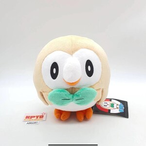 May include: A round, tan plush toy shaped like an owl with a white face, large black eyes, a small orange beak, and a green bowtie. The plush has orange feet and a black tag with a character illustration and the Japanese text モクロー.