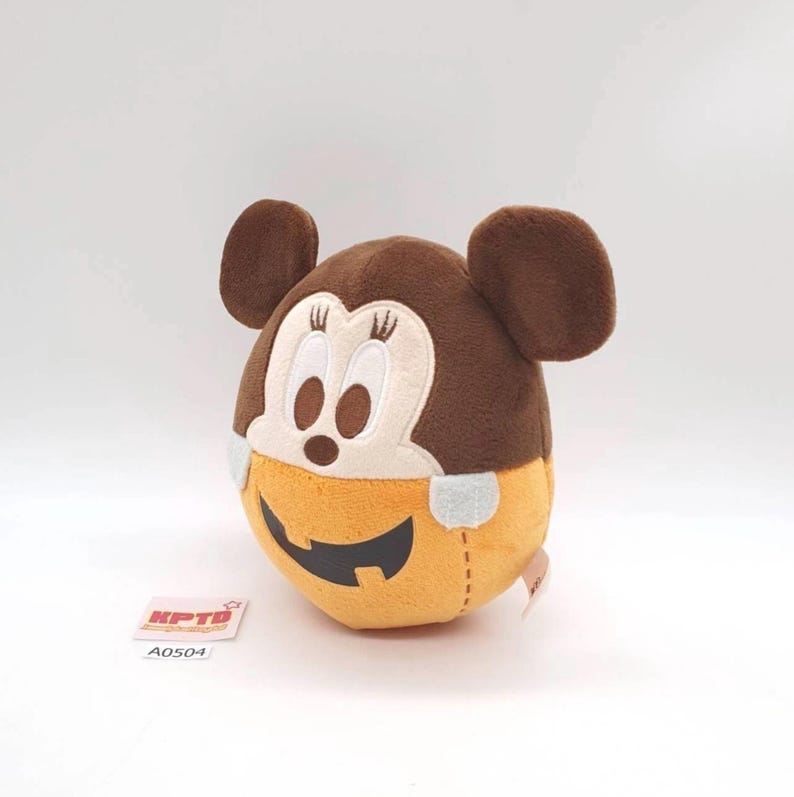 K&ouml;nnte beinhalten: Ein rundes Pl&uuml;schtier mit einem braunen Mickey-Maus-Kopf mit gestickten Gesichtsdetails und Wimpern, das auf einer orangefarbenen, k&uuml;rbisf&ouml;rmigen Basis mit einem schwarzen K&uuml;rbisgesicht sitzt. Ein kleines wei&szlig;es Etikett mit KPTD und A0504 ist sichtbar.