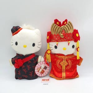 May include: Two plush Hello Kitty dolls dressed in traditional Chinese wedding attire. One wears a black and red outfit, the other a red wedding dress with a gold headdress. A tag with the text "KPTB" and "C0620" is visible.