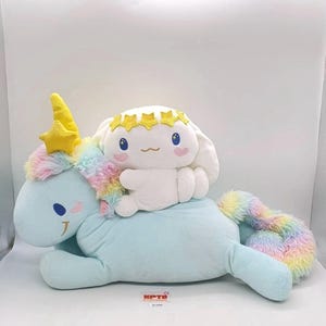 Puede incluir: Un juego de peluches con un unicornio azul claro con melena y cola arcoíris, y un personaje blanco con una corona de estrellas. El unicornio tiene un cuerno y una estrella amarillos. El personaje blanco está montando el unicornio.