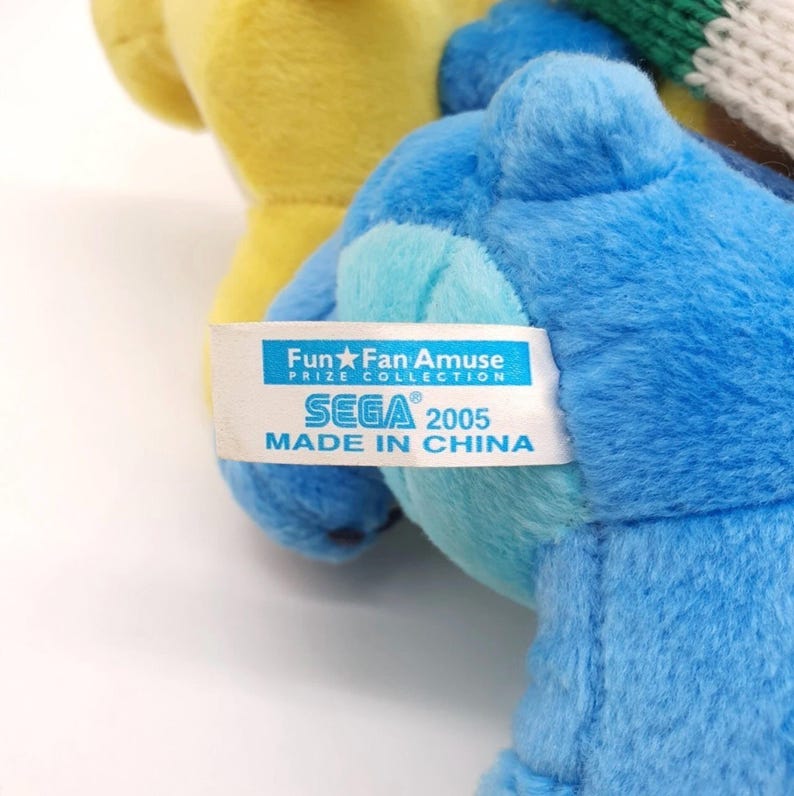 Puede incluir: Un peluche azul con una etiqueta de tela blanca que muestra el texto Fun Fan Amuse PRIZE COLLECTION SEGA 2005 MADE IN CHINA. El juguete tiene una textura suave y esponjosa y est&aacute; colocado junto a un peluche amarillo y una tela tejida verde y blanca.