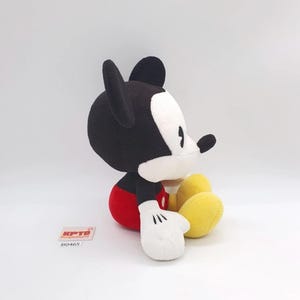K&ouml;nnte beinhalten: Ein Pl&uuml;schtier der Figur Mickey Maus in sitzender Position mit schwarzem Kopf und Ohren, wei&szlig;em Gesicht, roter Hose, wei&szlig;en Handschuhen und gelben Schuhen. Ein kleines wei&szlig;es rechteckiges Etikett mit dem roten Text KPTD und B0465 ist sichtbar.