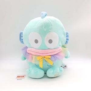May include: A plush toy in the shape of a cartoon creature, primarily light blue with large white eyes and gray pupils. It has blue ear-like appendages and a small crest. A pink bib and rainbow-colored tulle scarf are around its neck, with a yellow bow.
