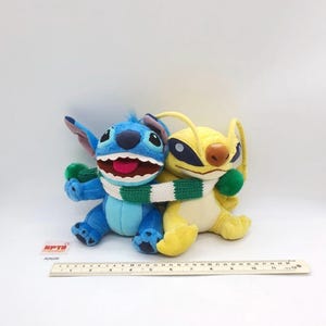 Puede incluir: Dos peluches, uno azul y otro amarillo, sentados uno al lado del otro y envueltos en una bufanda de punto a rayas verdes y blancas. Una regla colocada delante muestra que los juguetes miden aproximadamente 30 cm de ancho total en la base.