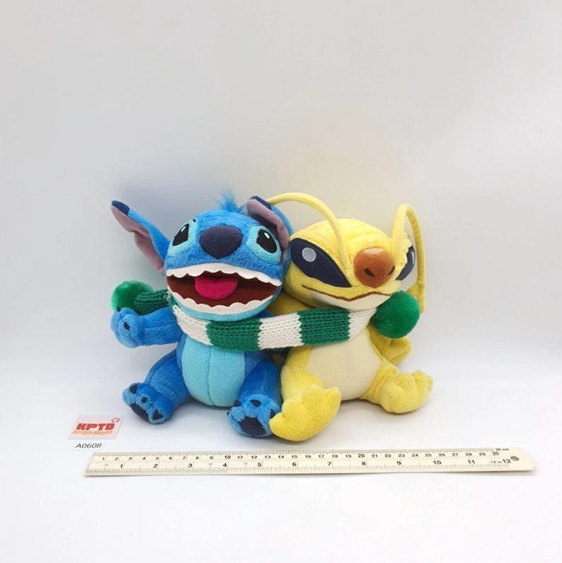 Puede incluir: Dos peluches, uno azul y otro amarillo, sentados uno al lado del otro y envueltos en una bufanda de punto a rayas verdes y blancas. Una regla colocada delante muestra que los juguetes miden aproximadamente 30 cm de ancho total en la base.