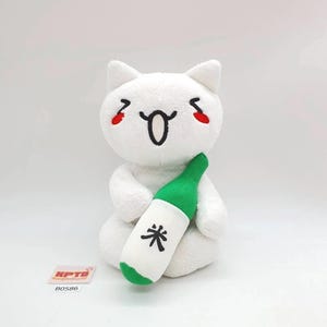 以下が含まれることがあります： 笑顔と赤い頬のマークが付いた白いぬいぐるみ猫のおもちゃで、日本語のテキストが入った緑と白のボトルを持っています。おもちゃには、ステッチされた黒い口と目があります。ボトルは緑色のキャップと黒い文字の白いラベルが付いています。