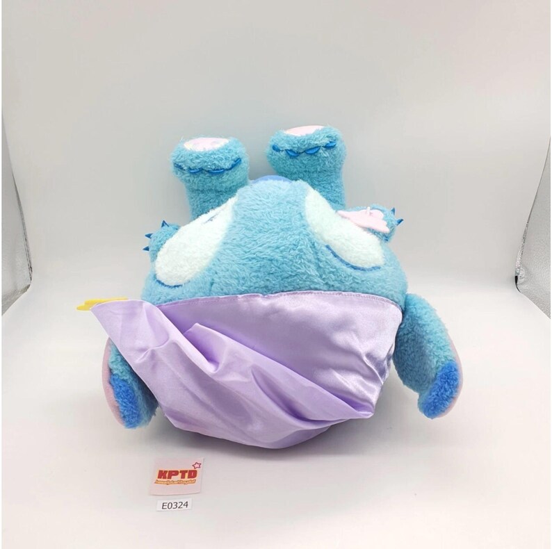 May include: A plush toy in the shape of a blue creature with large ears and a lavender satin cape. The toy has white accents and is lying on its back. The tag reads "KPTD" and "E0324".