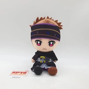 Puede incluir: Un muñeco de peluche con cabello castaño en punta, una cinta para la cabeza morada y negra y grandes ojos rosas. El muñeco viste un conjunto negro con un cinturón plateado y un emblema amarillo y negro. Se ve una etiqueta blanca con KPTD H0322.
