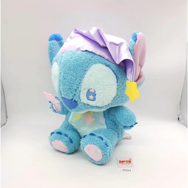 May include: A plush Stitch doll in a seated position, featuring a light blue body, large white eyes, and a blue nose. It wears a lavender nightcap and holds a pink item. A yellow star-shaped charm hangs from its ear. The doll has pink accents on its ears and feet.