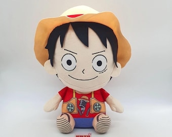 May include: A plush doll of a character from the anime series One Piece. The doll has a tan hat, black hair, and a red shirt with brown suspenders. The doll is sitting with its legs bent and has a smiling expression.