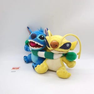 Puede incluir: Dos peluches, uno azul y otro amarillo, unidos por una bufanda de punto verde y blanca. El peluche azul tiene orejas grandes, una boca ancha con dientes visibles y una lengua rosa. El amarillo presenta antenas largas, parches oscuros y nariz marr&oacute;n.
