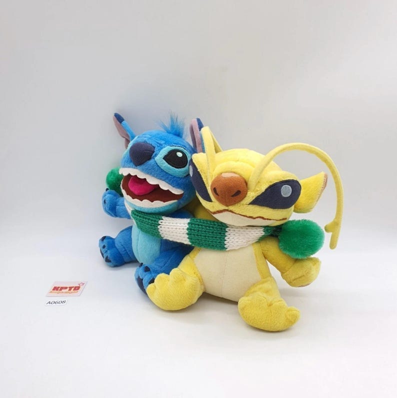Puede incluir: Dos peluches, uno azul y otro amarillo, unidos por una bufanda de punto verde y blanca. El peluche azul tiene orejas grandes, una boca ancha con dientes visibles y una lengua rosa. El amarillo presenta antenas largas, parches oscuros y nariz marr&oacute;n.