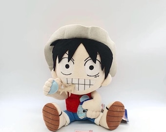 May include: A plush toy of the anime character Monkey D. Luffy from One Piece. He is wearing a red shirt, blue shorts, and a straw hat. He is holding a blue and white plush fish in his right hand and a blue and white plush starfish in his left hand. The toy is sitting on a white surface. The text "KPTD 30572" is visible on the bottom of the image.