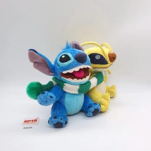 Puede incluir: Dos peluches que representan un personaje azul con orejas grandes y una amplia sonrisa que lleva una bufanda de punto a rayas verdes y blancas, junto a un personaje amarillo con parches oscuros en los ojos y antenas. Una etiqueta KPTD A0608.