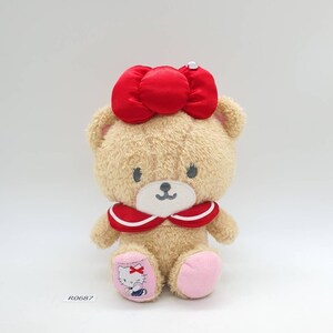 Può includere: Un orsacchiotto di peluche con un fiocco rosso sulla testa e un colletto rosso e bianco. L'orso ha una faccia sorridente e zampe rosa. Il piede sinistro presenta un design di Hello Kitty. L'etichetta recita R0687.