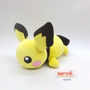 May include: A yellow plush toy with black accents on the ears and back. The toy has a pink cheek and a black eye. A small white tag with the text "KPTD" and the number "N0566" is visible.