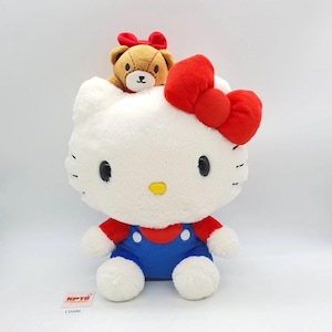 May include: A large white plush Hello Kitty doll with a red bow, red shirt, and blue overalls. A small brown teddy bear with a red bow sits on top of the doll's head. The doll has black eyes and a yellow nose.