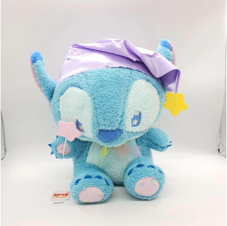 May include: A plush Stitch toy in a seated position, featuring a light blue body, large white eyes, and a blue nose. It wears a lavender sleep cap and has a yellow star and pink star-shaped accessories. The toy has a tag with the text "KPTB E0324".