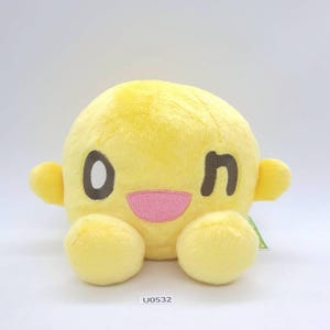 Puede incluir: Un peluche amarillo con forma de personaje sonriente con un guiño, una boca rosa sonriente y pequeños brazos y pies. El texto "U0532" es visible en la parte inferior. El peluche está sobre un fondo blanco.