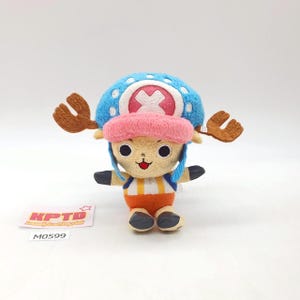以下が含まれることがあります： 青い帽子に白い十字、ピンクのつば、茶色の角を持つキャラクターのぬいぐるみ。 キャラクターは、タン色の顔、オレンジと白のストライプのシャツ、黒い手と足を持っています。 テキストの入った小さなタグが見えます。