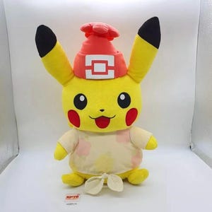 May include: A yellow Pikachu plush toy wearing a red beanie with a white square emblem and a cream-coloured shirt featuring a light pink and yellow floral pattern. The shirt is tied at the bottom with a knot. The plush has black-tipped ears and red cheeks.