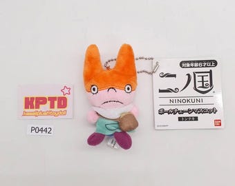 May include: A plush toy of a character from the video game Ni no Kuni. The toy has orange ears, a pink face, and is wearing a green shirt and purple shoes. It has a small brown bag and a metal chain.