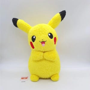 May include: A yellow Pikachu soft toy with black-tipped ears and red cheek patches. The toy has a smiling face with a pink tongue and is standing with its hands together. A small tag with text is visible at the bottom.