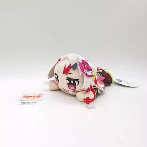 May include: A plush toy featuring a character with white hair, large dark eyes, and a small pink demon mask accessory on the side of the head. The figure has red accents, a patterned outfit, and is positioned horizontally. A small tag reads KPTD M00214.