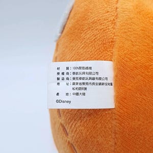 K&ouml;nnte beinhalten: Ein leuchtend orangefarbenes Pl&uuml;schtier mit einem wei&szlig;en Stoffetikett. Das Etikett enth&auml;lt schwarzen Text in chinesischen Schriftzeichen, der Materialzusammensetzung, Hersteller und Herkunft angibt, sowie das Disney-Logo unten.