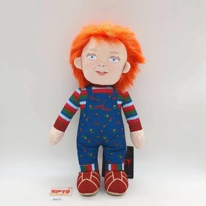 May include: A plush doll with bright orange hair, blue eyes, and freckles. It wears a long-sleeved shirt with horizontal multi-colored stripes and blue denim-style overalls featuring a small red Good Guys logo and a colorful pattern of various icons.