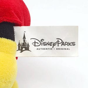K&ouml;nnte beinhalten: Ein Pl&uuml;schtier mit einem wei&szlig;en Stoffetikett, das den schwarzen Schriftzug Disney Parks Authentic Original und eine Schlossgrafik zeigt. Das Spielzeug hat einen oberen Teil aus weichem rotem Stoff, einen schwarzen Mittelteil und einen gelben Unterteil.