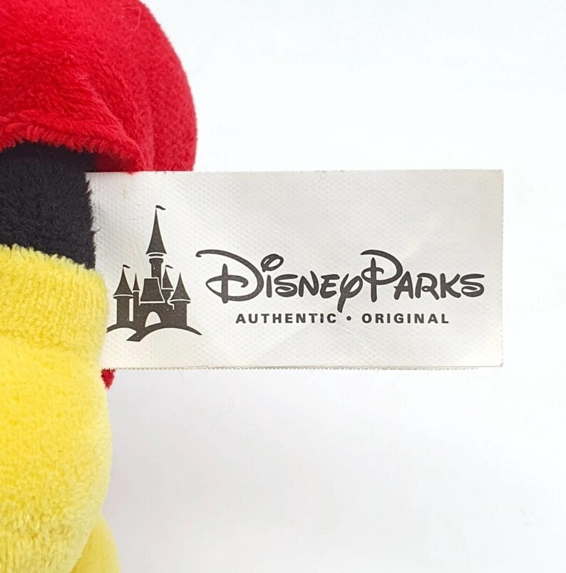 K&ouml;nnte beinhalten: Ein Pl&uuml;schtier mit einem wei&szlig;en Stoffetikett, das den schwarzen Schriftzug Disney Parks Authentic Original und eine Schlossgrafik zeigt. Das Spielzeug hat einen oberen Teil aus weichem rotem Stoff, einen schwarzen Mittelteil und einen gelben Unterteil.