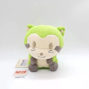 May include: A plush toy shaped like a cat with bright lime green fur, a cream-coloured face, and dark grey paws and ear accents. The toy features embroidered eyes, a small nose, and whiskers. A small tag with the text KPTD M00176 is visible nearby.