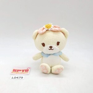 May include: A cream-colored plush bear with a flower headband and a blue collar. The bear has brown eyes and a small brown nose. The bear's paws have pink accents. A tag with the text "KPTD" and the number "L0479" is in the lower left corner.
