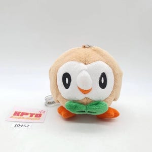 May include: A round, tan plush toy featuring a white facial disc, black oval eyes, a small orange beak, and a green bowtie-shaped feature at the base. The plush has orange feet and a small metal ball chain attached to the top. A tag reads KPTD I0452.