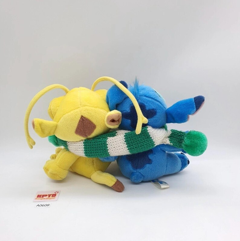 Puede incluir: Dos peluches, uno amarillo y otro azul, se abrazan mientras llevan una bufanda de punto a rayas verdes y blancas compartida. El juguete amarillo tiene antenas largas y el azul tiene orejas grandes. Una peque&ntilde;a etiqueta blanca dice KPTD A0608.