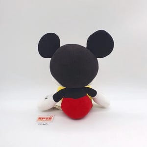 K&ouml;nnte beinhalten: Ein Pl&uuml;schtier der Figur Mickey Mouse mit gro&szlig;en, schwarzen, abgerundeten Ohren, einem schwarzen Kopf, wei&szlig;en Handschuhen und einer leuchtend roten Hose. Ein kleines rechteckiges Etikett mit dem Text KPTD und B0465 befindet sich an der Basis.