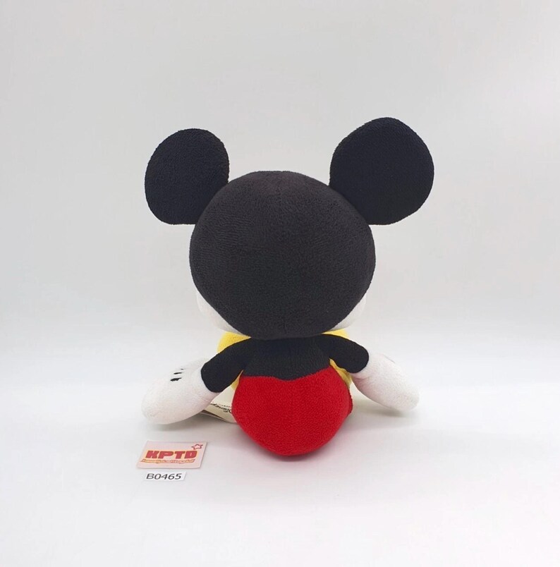 K&ouml;nnte beinhalten: Ein Pl&uuml;schtier der Figur Mickey Mouse mit gro&szlig;en, schwarzen, abgerundeten Ohren, einem schwarzen Kopf, wei&szlig;en Handschuhen und einer leuchtend roten Hose. Ein kleines rechteckiges Etikett mit dem Text KPTD und B0465 befindet sich an der Basis.