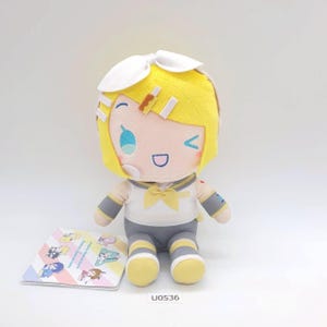 Może przedstawiać: Pluszowa lalka z żółtymi włosami i białą kokardą. Lalka ma na sobie biało-szary strój z żółtą kokardą. Obok lalki znajduje się mała karta z postaciami z kreskówek. Kod produktu "U0536" jest wydrukowany na dole.