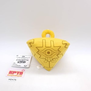 Könnte beinhalten: Gelbes Plüschtier in Dreiecksform mit detailliertem Augendesign und geometrischen Mustern. Es hat eine kleine Schlaufe zum Aufhängen. Ein Etikett mit japanischem Text ist angebracht. Wahrscheinlich ein Sammlerstück oder ein Charakterartikel.