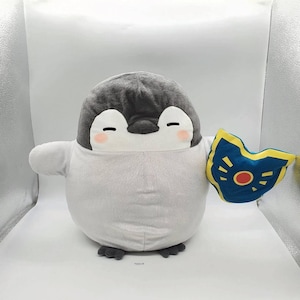 May include: A plush penguin toy with a gray head and white body, holding a blue and yellow shield. The penguin has closed eyes and rosy cheeks, giving it a friendly appearance. The shield has a red circle in the center.