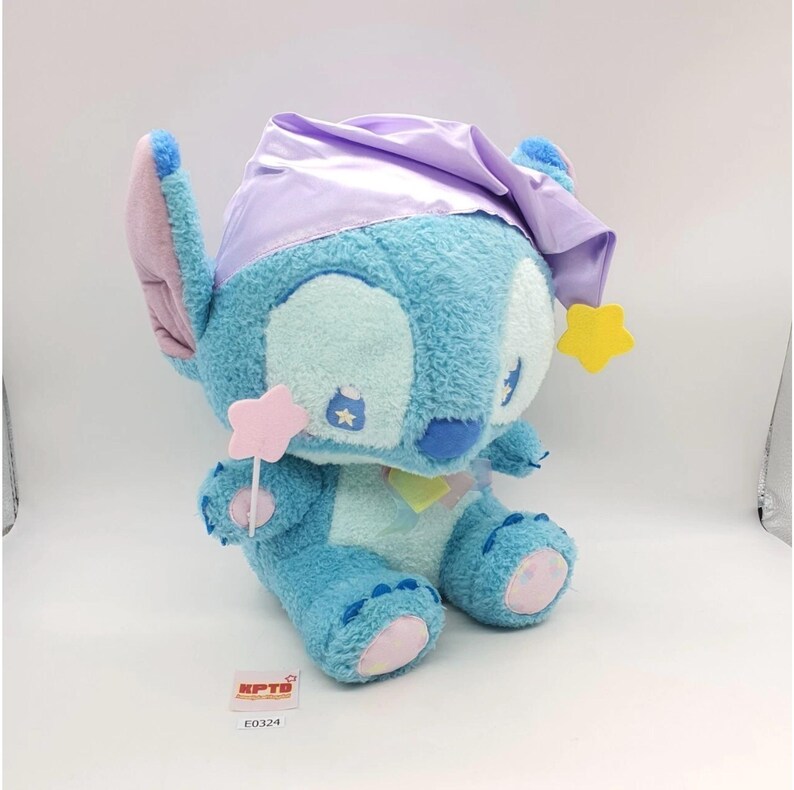 May include: A plush Stitch toy in a seated position, featuring a light blue body, large ears, and a lavender nightcap. The toy holds a pink star wand and has yellow star accents. The toy has a tag that reads "KPTB" and "E0324".