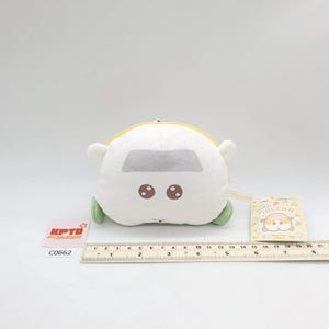 Peut inclure: Peluche blanche en forme de voiture avec une fenêtre grise, des yeux marron et des roues vertes. Elle possède une fermeture éclair jaune et de petites oreilles blanches. Une étiquette avec le texte "MOLCAR" est attachée.
