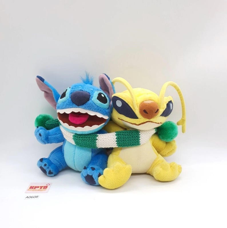 Puede incluir: Dos peluches, un personaje azul de Stitch con una amplia sonrisa y un personaje alien&iacute;gena amarillo, est&aacute;n unidos por una bufanda de punto a rayas verdes y blancas. El personaje azul tiene orejas grandes y el amarillo tiene dos antenas largas.