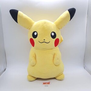May include: A large, plush Pikachu toy in a standing pose. The toy is primarily yellow with black-tipped ears, black eyes, red cheek circles, and a small, smiling mouth. The toy has a tag with the text "KPTD" and "M0500".