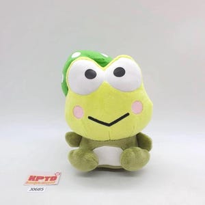 May include: A plush frog toy in shades of green and yellow, with large black eyes and pink cheeks. The frog wears a green hat with white polka dots. The toy has a smiling expression and is sitting upright. A tag with the text "KPTD" and "J0685" is visible.