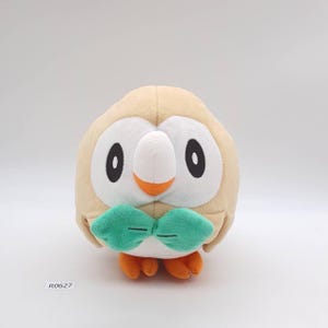 May include: A plush toy owl with a tan body, white face, and large black eyes. It has an orange beak, green bow tie, and orange feet. The toy is designed to resemble a cartoon character.
