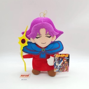 May include: A plush doll featuring a character with bright purple hair, wearing a red tunic with a blue collar and brown shoes. The doll holds a yellow star-shaped staff and has an attached tag with Japanese text and an illustration of characters.